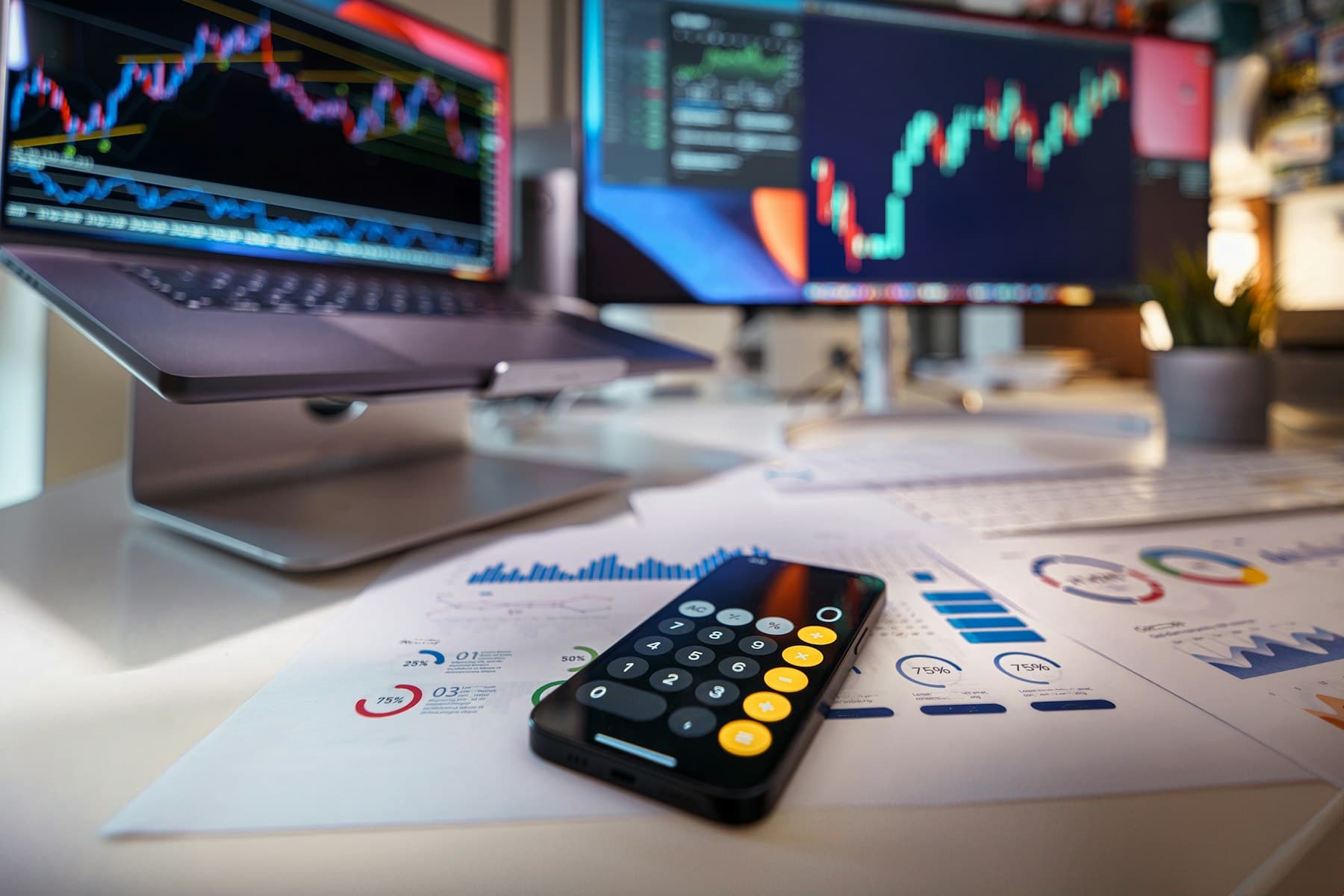 Calculator on a desk in front of monitors with financial charts